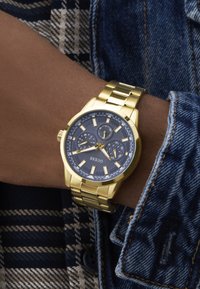 Złoty zegarek ze stali nierdzewnej z granatową teksturowaną tarczą, świecącymi znacznikami godzin, trzema subtarcza oraz bransoletą siateczkową, noszony na nadgarstku.