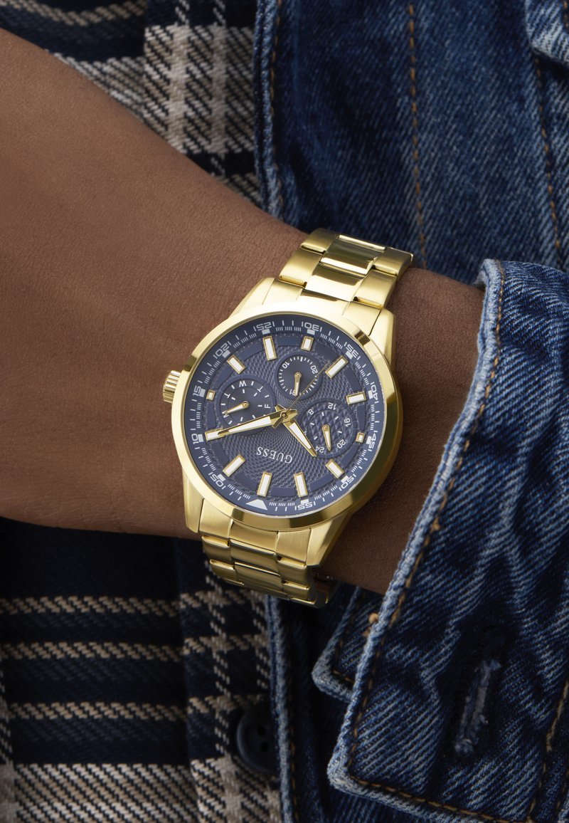 Złoty zegarek ze stali nierdzewnej z granatową teksturowaną tarczą, świecącymi znacznikami godzin, trzema subtarcza oraz bransoletą siateczkową, noszony na nadgarstku.