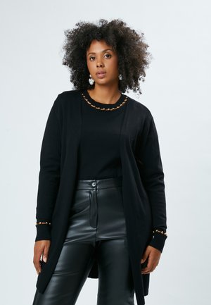 Cardigan nero con una texture liscia, caratterizzato da un collo rotondo decorato con perline dorate e polsini a coste, indossato sopra una maglietta nera e pantaloni di pelle.
