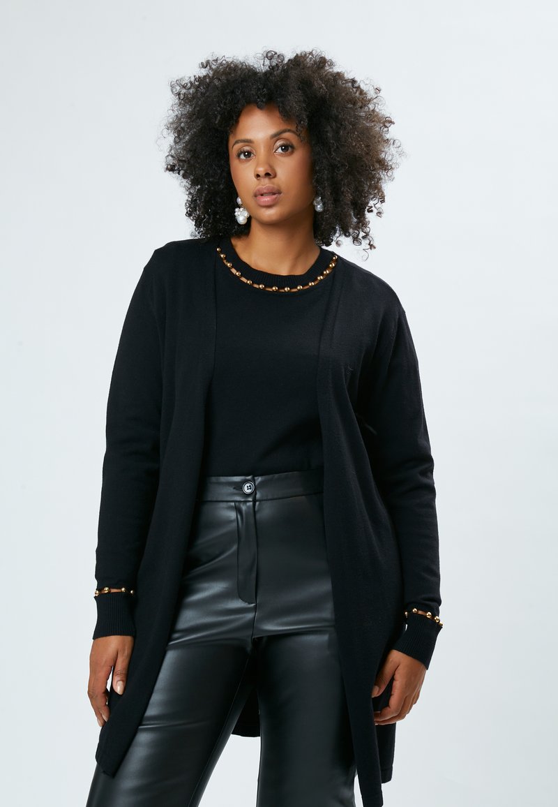 Gilet noir au toucher lisse, avec un col rond orné de perles dorées et des poignets côtelés, porté sur un haut noir et un pantalon en cuir.