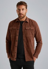 Bruin geruit shirt met een knoopsluiting aan de voorkant, twee borstzakken en een lichte, gestructureerde stof. Draag het over een zwart shirt.