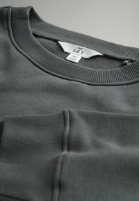 Sweatshirt à col ras du cou gris foncé avec col et poignets côtelés, affichant une étiquette blanche de taille portant la mention "THE SET" et "M Fabriqué au Sri Lanka."