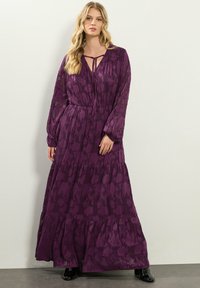 Długa, warstwowa sukienka maxi w głębokim fioletowym materiale w kwiatowy wzór. Posiada długie rękawy, luźny krój oraz detal w postaci wiązania przy dekolcie.