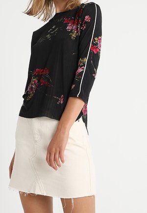 Femme portant un haut noir à fleurs à manches trois-quarts avec des bandes blanches sur les côtés et une mini-jupe en jean blanche à ourlet effiloché.