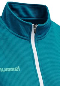 Giacca zip-up teal realizzata in tessuto testurizzato, con colletto alto e zip bianca. Il logo è stampato in azzurro chiaro sul lato sinistro.