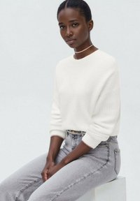 Bel bela pletena pulover s rebrastimi rokavi, v paru z visokim pasom svetlo sivih kavbojk, sedeč ob svetli podlagi.