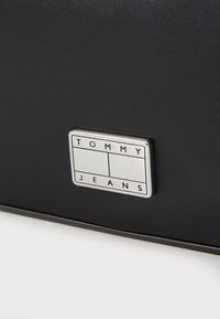 Svart läderhandväska med en silverfärgad rektangulär logoplåt med "TOMMY JEANS" ingraverat. Slät yta med rena linjer.