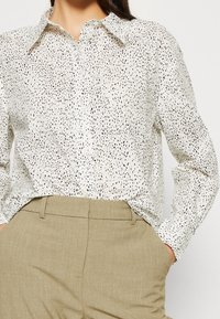Knoop-sluiting overhemd in crèmekleur met een zwart gevlekt patroon, met een kraag en lange mouwen. Gepaard met lichtbeige broeken.