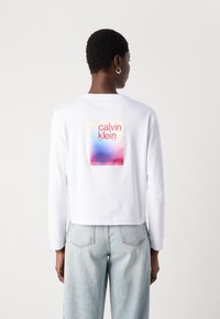 T-shirt bianco a maniche lunghe con un logo quadrato colorato sulla schiena, con testo in rosso, blu e rosa. Ha una vestibilità morbida.