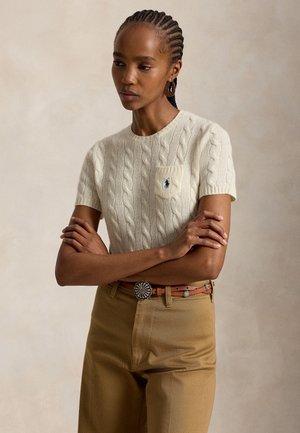 Femme aux cheveux tressés portant un pull court blanc en maille torsadée et un pantalon beige avec une ceinture marron, bras croisés, fond neutre.