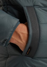 Manica di giacca imbottita in un verde scuro con una texture liscia e lucida. Presenta un polsino elastico nero e un'etichetta del logo visibile.
