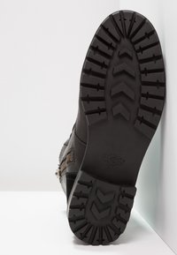 Botín negro hasta el tobillo con una suela de goma texturizada con un patrón en zigzag y un detalle de cremallera lateral para facilitar su uso. Logo de UGG visible.