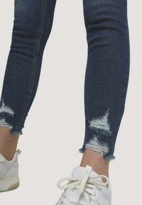 Mörkblå jeans i skinny-modell med slitna knädetaljer och fransad fåll, kombinerade med vita sneakers. Slät textur och figurnära design.