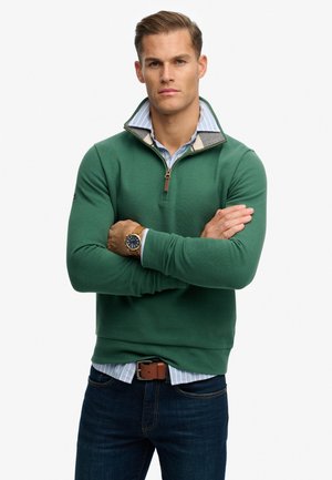 Mand med korslagte arme iført en grøn quarter-zip trøje, blåstribet skjorte, mørke jeans, brunt bælte og armbåndsur mod en hvid baggrund.