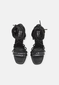 Sandalias de tacón alto negras con tiras finas, punta cuadrada y una textura suave. Los acentos transparentes en las correas añaden detalle.