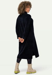 Manteau long noir et duveteux avec un col montant et des fentes sur les côtés. Porté par-dessus des leggings noirs et des bottes beiges avec des accents verts.