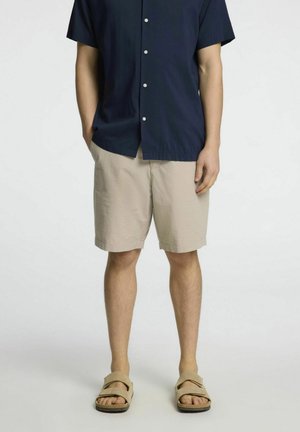 Homme portant un short beige, une chemise à manches courtes bleu marine à boutons et des sandales beige, debout devant un fond uni.