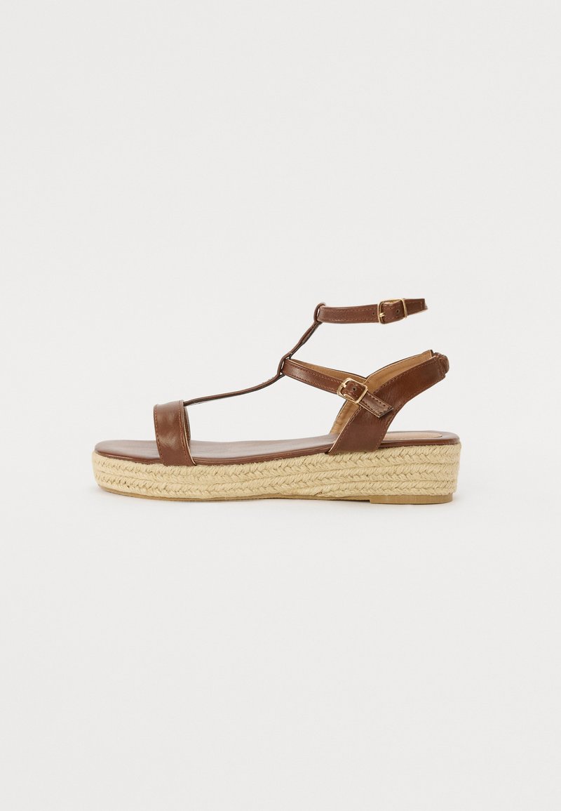 Brown leather sandal with a T-strap design, adjustable ankle straps, and a braided jute wedge sole. Flat profile and smooth finish.