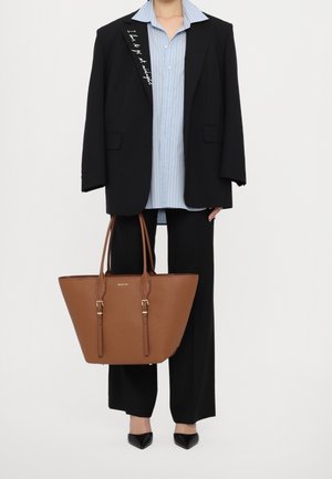 Personne portant un blazer noir et un pantalon avec une chemise à rayures bleues claires, tenant un grand sac cabas en cuir marron et portant des chaussures noires à bouts pointus.