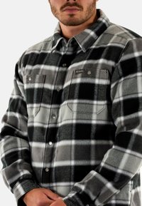 Camicia in flanella a scacchi grigia e nera con due tasche sul petto e chiusura a bottoni a pressione, caratterizzata da un tessuto testurizzato e morbido. Dettaglio del logo sulla tasca sinistra.