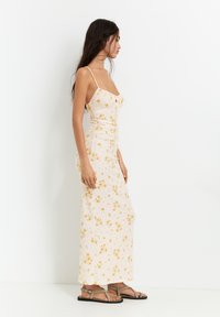 Rochie maxi florală în crem cu flori galbene, având bretele subțiri, detalii pliate și o siluetă ajustată. Asortată cu sandale plate.