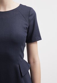 Kvinna iförd en mörkblå texturerad kortärmad klänning med plisserad detalj på sidan, mot en enfärgad ljus bakgrund.
