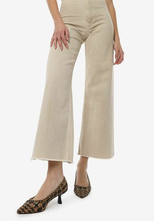 Beige high-waisted wide-leg jeans with frayed hems, paired with black and orange patterned pointed-toe heels on a model standing.