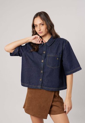 Vrouw draagt een losse donkerdenim blouse met korte mouwen en een bruine wikkelrok, poseert met één hand bij haar gezicht.