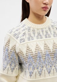 Mulher vestindo um suéter de malha creme com padrões geométricos texturizados em bege e azul claro no peito e nas mangas.