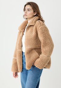 Beżowa kurtka ze sztucznego futerka z wysokim kołnierzem, zamkiem błyskawicznym z przodu i luźnym krojem. Posiada fakturowaną tkaninę i boczne kieszenie, zestawiona z niebieskimi dżinsami.