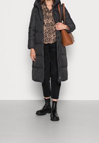 Svart puffjacka med huva, mönstrad brun långärmad topp, svarta kortbyxor och svarta ankelskot. Brun handväska. Slät textur.