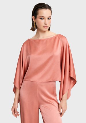 Žena s krátkymi tmavými vlasmi, ktorá má zlaté náušnice a nosí voľný, lesklý koralovo ružový saténový top a zodpovedajúce nohavice na neutrálnom svetlom pozadí.