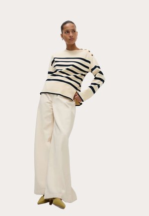 Maglione a righe crema e blu navy con bottoni dorati, abbinato a pantaloni crema a gamba larga e scarpe gialle con punta a punte su sfondo neutro.