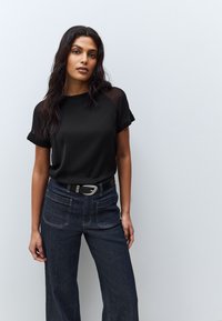 Femme aux longs cheveux foncés portant un haut noir à manches courtes et un jean sombre taille haute avec poches avant et ceinture, debout contre un mur uni.