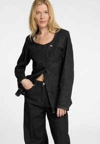 Svart denimjacka med fyrkantig halsringning, långa ärmar och diagonalt framknäppning. Tyget har en lätt texturerad yta och en dekorativ brosch.