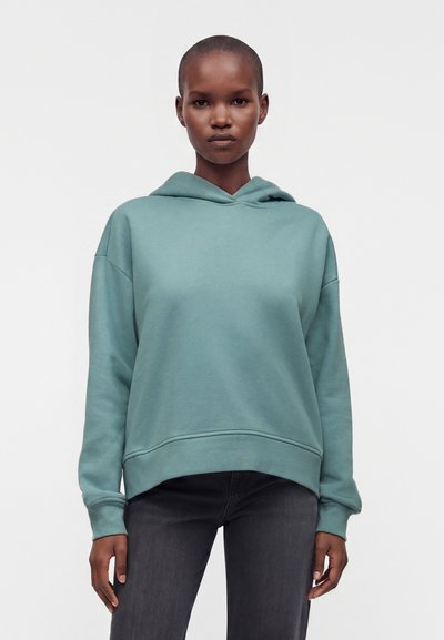 Personne aux cheveux courts porte un sweat à capuche ample bleu canard et un jean foncé, debout face à un fond uni clair.
