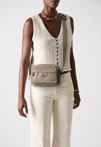 Borsa a tracolla marrone con chiusura a zip, tessuto texture e dettagli con logo. Tracolla regolabile e tasca frontale. Modello che indossa un maglione lavorato a maglia e jeans chiari.
