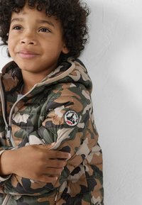 Camouflage pufferjack in groen, bruin en beige met grijze accenten. Voorzien van een rits aan de voorkant en een ronde logopatch op de linkerarm.