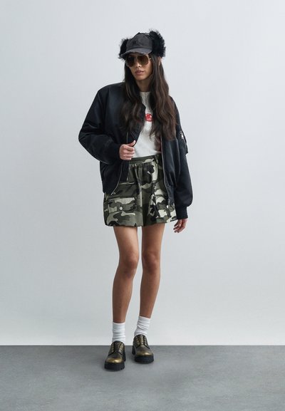 Schwarze Bomberjacke über einem weißen T-Shirt, Camouflage-Shorts, weiße Socken und schwarz-goldene Schuhe. Die Kappe ist mit schwarzem Flausch verziert.