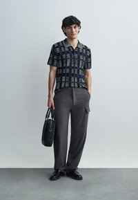 Short-sleeve, collared polo shirt in a patterned navy and green design, paired with gray, wide-leg trousers and a black bag. Smooth texture.