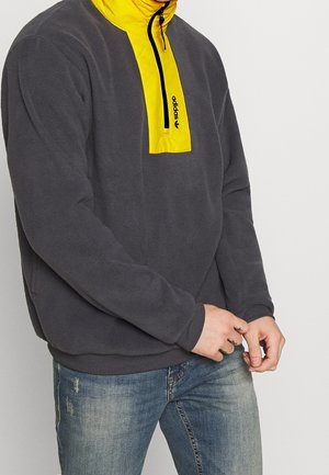 Person trägt dunkelgrauen Fleecepullover mit gelbem Reißverschluss am Kragen und blaue, abgenutzte Jeans, die Hände vor dem Körper verschränkt.