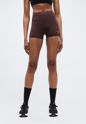 Persoană slabă purtând un top scurt negru, pantaloni scurți atletici maro, talie înaltă, șosete negre până la mijlocul gambei și pantofi de alergare negri, stând drept.