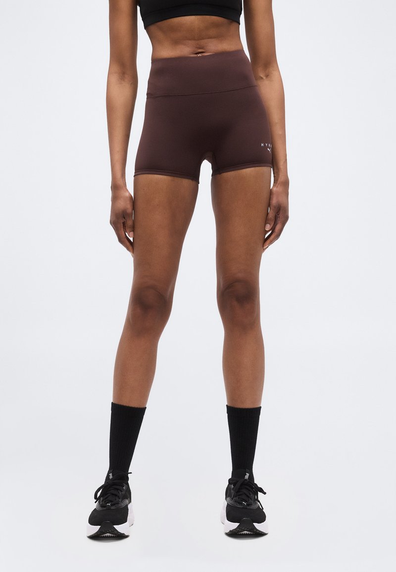 Λεπτό άτομο που φοράει μαύρο cropped τοπ, καφέ αθλητική σορτς με ψηλή μέση, μαύρες κάλτσες μέχρι τη μέση της γάμπας και μαύρα παπούτσια τρεξίματος, στέκεται όρθιο.