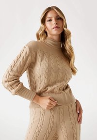 Beiger Kabelstrickpullover mit hohem Kragen und gerippten Bündchen, der mit aufwendigen strukturierten Mustern versehen ist, kombiniert mit passenden Kabelstrickhosen.