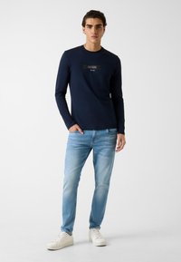 Granatowa koszulka z długim rękawem z czarnym i srebrnym logo „GUESS”, w połączeniu z jasnoniebieskimi dżinsami i białymi trampkami, na neutralnym tle.
