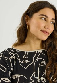 Haut noir avec des motifs abstraits de visages beiges. Modèle portant des boucles d'oreilles créoles dorées, avec des cheveux bruns ondulés sur un fond clair.