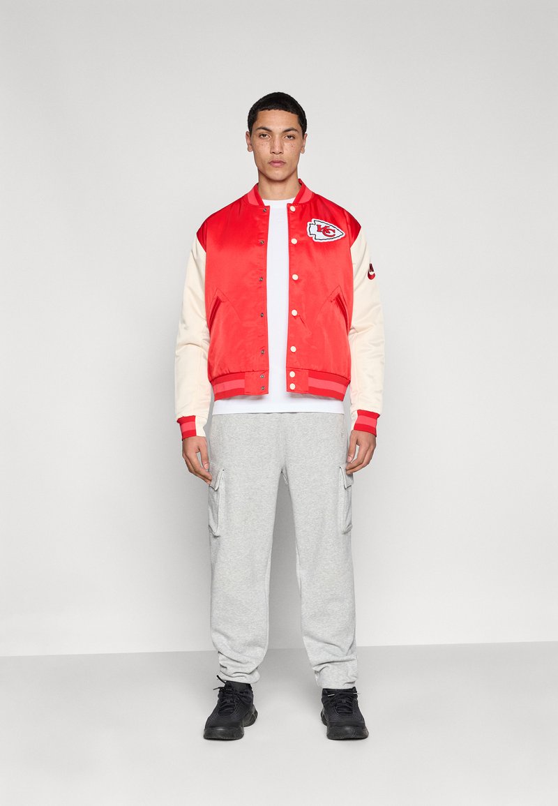 Rote Satin-Collegejacke mit cremefarbenen Ärmeln, verziert mit einem Logo, kombiniert mit grauen Jogginghosen und schwarzen Schuhen; Modell steht in einer neutralen Umgebung.