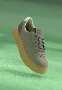 Sneaker din suede gri deschis, cu vârful perforat, șireturi gri și o talpă groasă galbenă. Dispune de un logo pe toc căptușit.