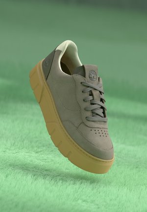 Light gray suede sneaker with perforated toe, gray laces, and a thick yellow outsole. Features a logo on the padded heel.