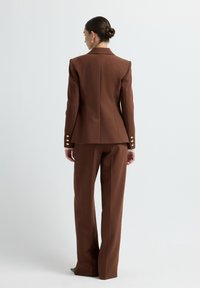 Blazer marrone su misura con spalle ben definite, abbottonatura sul retro e dettagli dorati. Abbinato a pantaloni a vita alta e gamba larga.